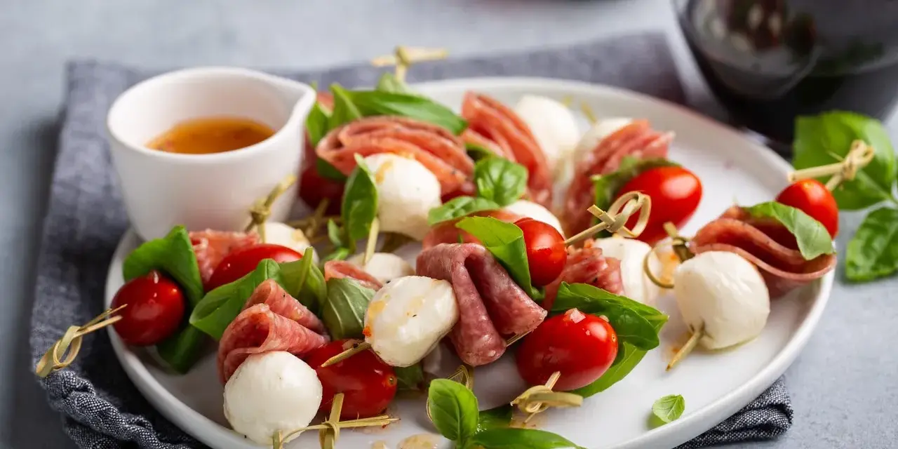 Pyszne keto przekąski na imprezę: szaszłyki z mozzarellą, pomidorkami i salami, ozdobione bazylią.