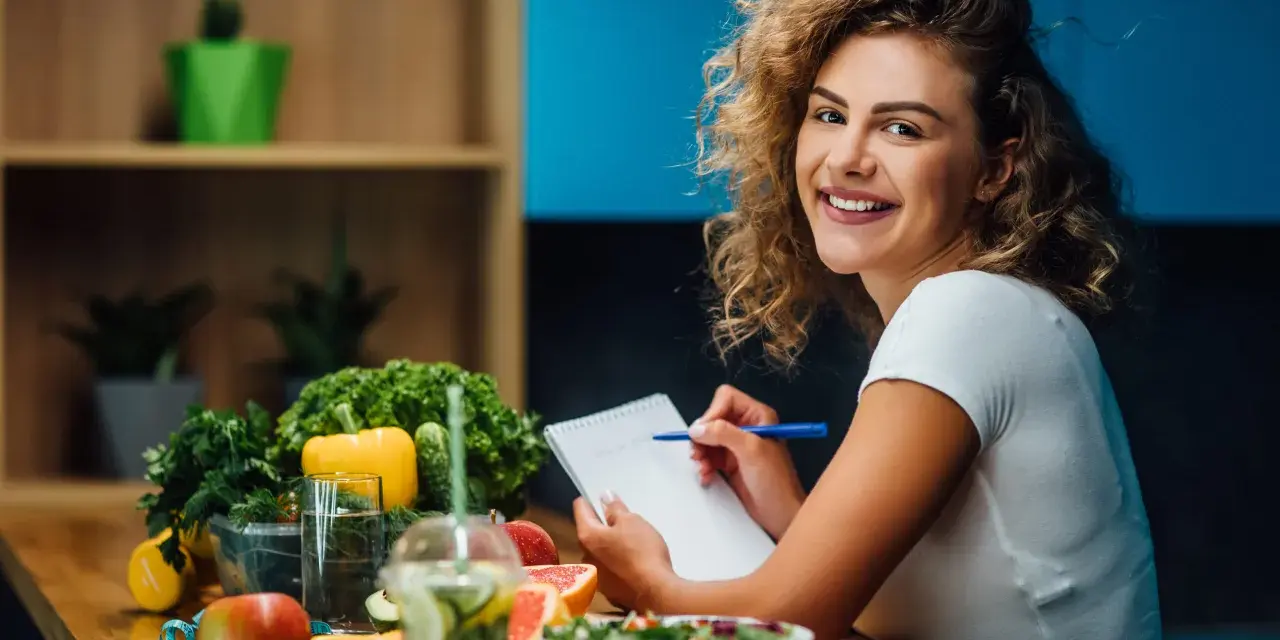 Uśmiechnięta kobieta notuje w zeszycie. Na stole świeże owoce i warzywa, idealne do diety 1500 kcal. Efekty po miesiącu będą widoczne!