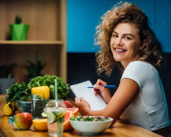 Uśmiechnięta kobieta notuje w zeszycie. Na stole świeże owoce i warzywa, idealne do diety 1500 kcal. Efekty po miesiącu będą widoczne!