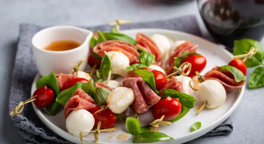 Pyszne keto przekąski na imprezę: szaszłyki z mozzarellą, pomidorkami i salami, ozdobione bazylią.