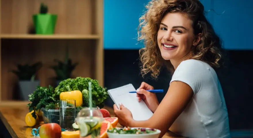 Uśmiechnięta kobieta notuje w zeszycie. Na stole świeże owoce i warzywa, idealne do diety 1500 kcal. Efekty po miesiącu będą widoczne!
